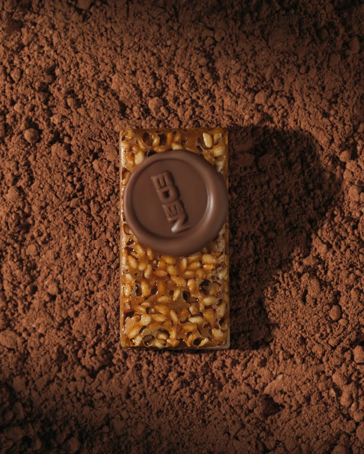 A nut brittle bar topped with a stamped chocolate disc, half buried in cocoa.