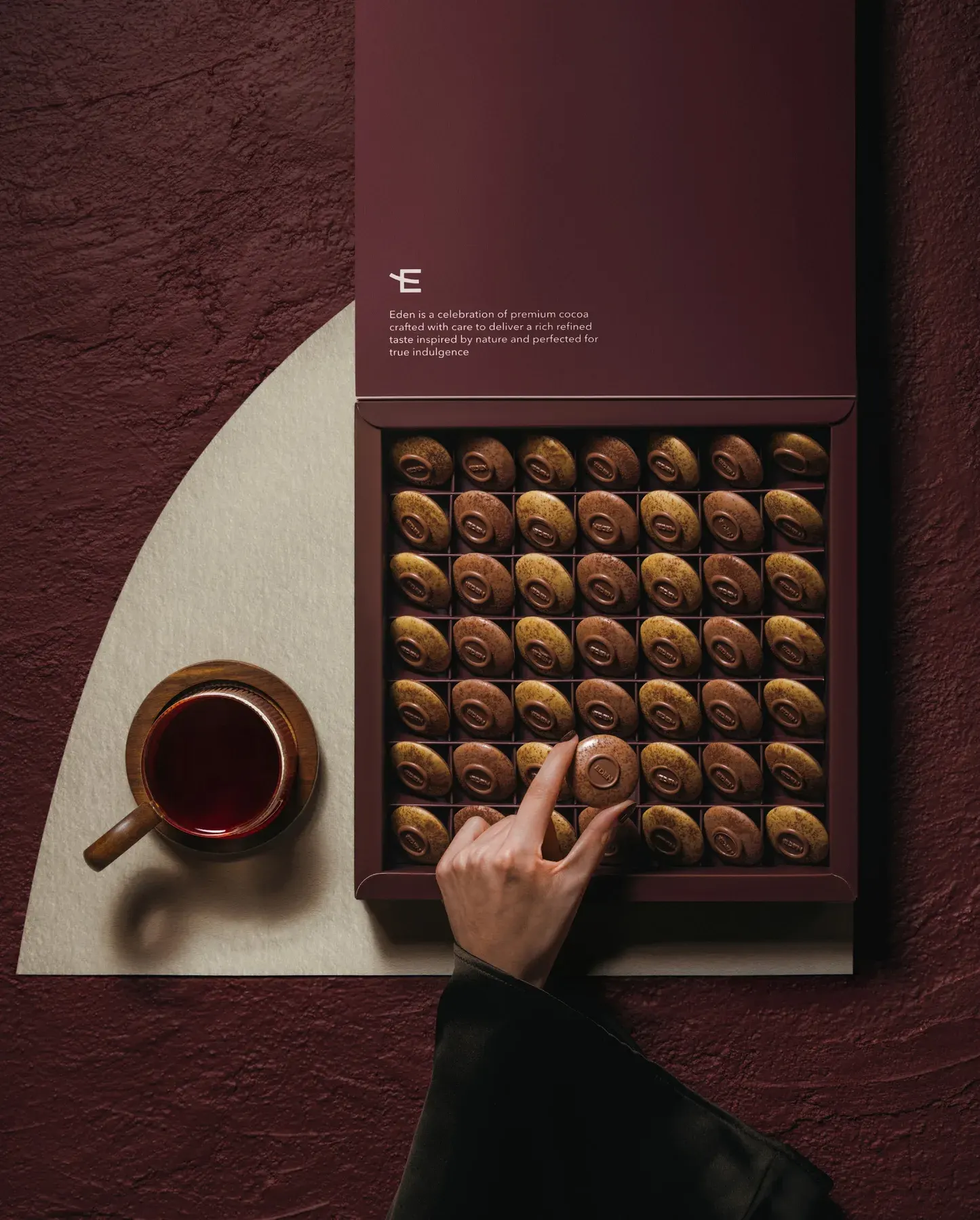 An open box of pralines beside a cup, a hand reaching for one piece.