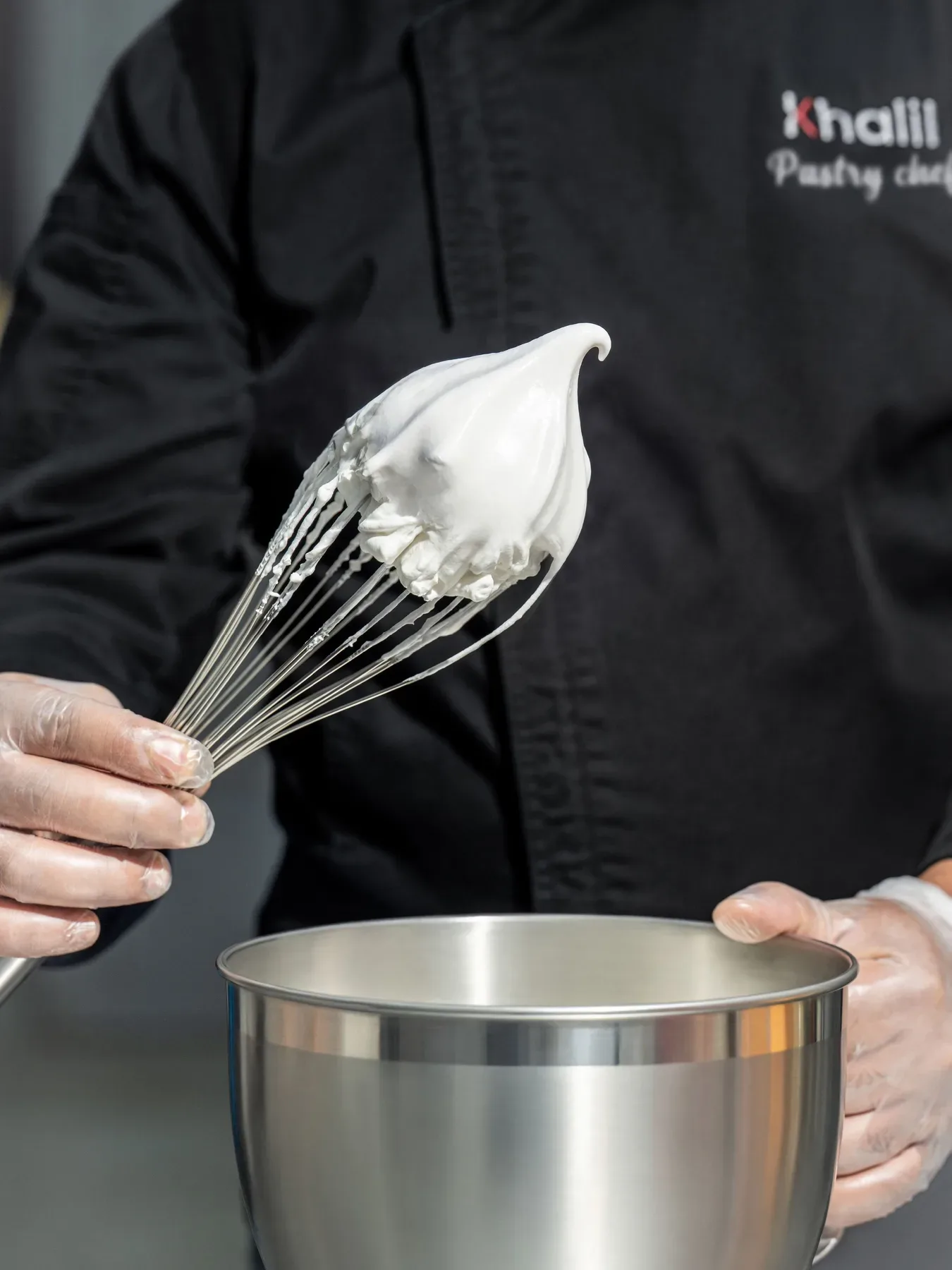 Whipped cream lifted on a whisk by a chef in a black Khalil jacket.