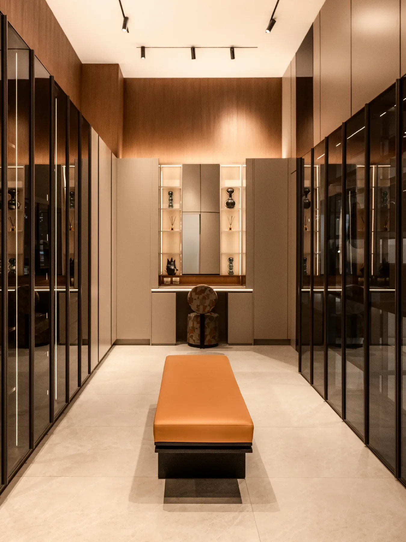 A walk in closet corridor with smoked glass doors and a tan leather bench.