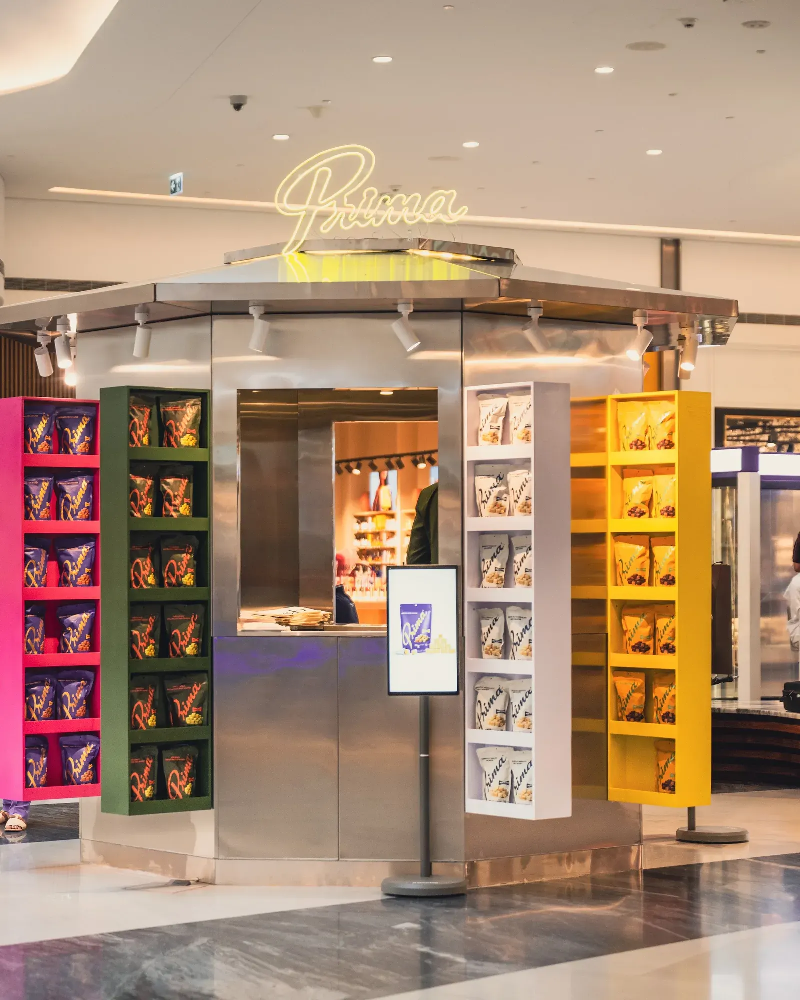 The Prima launch pop-up, a steel booth with a shelf in each flavour colour and the neon wordmark above.