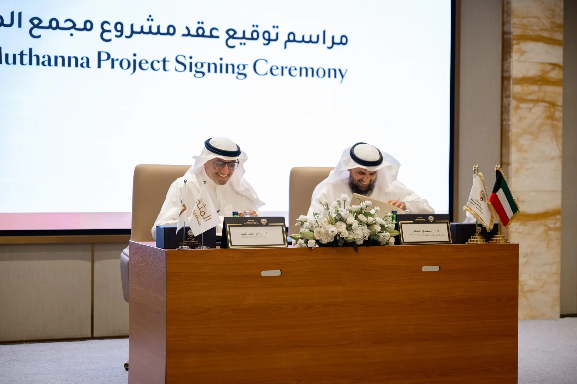 Two officials sign at a table under a screen reading "Al Muthanna Project Signing Ceremony", with Al Muthanna, Ministry of Finance and Kuwaiti desk flags.