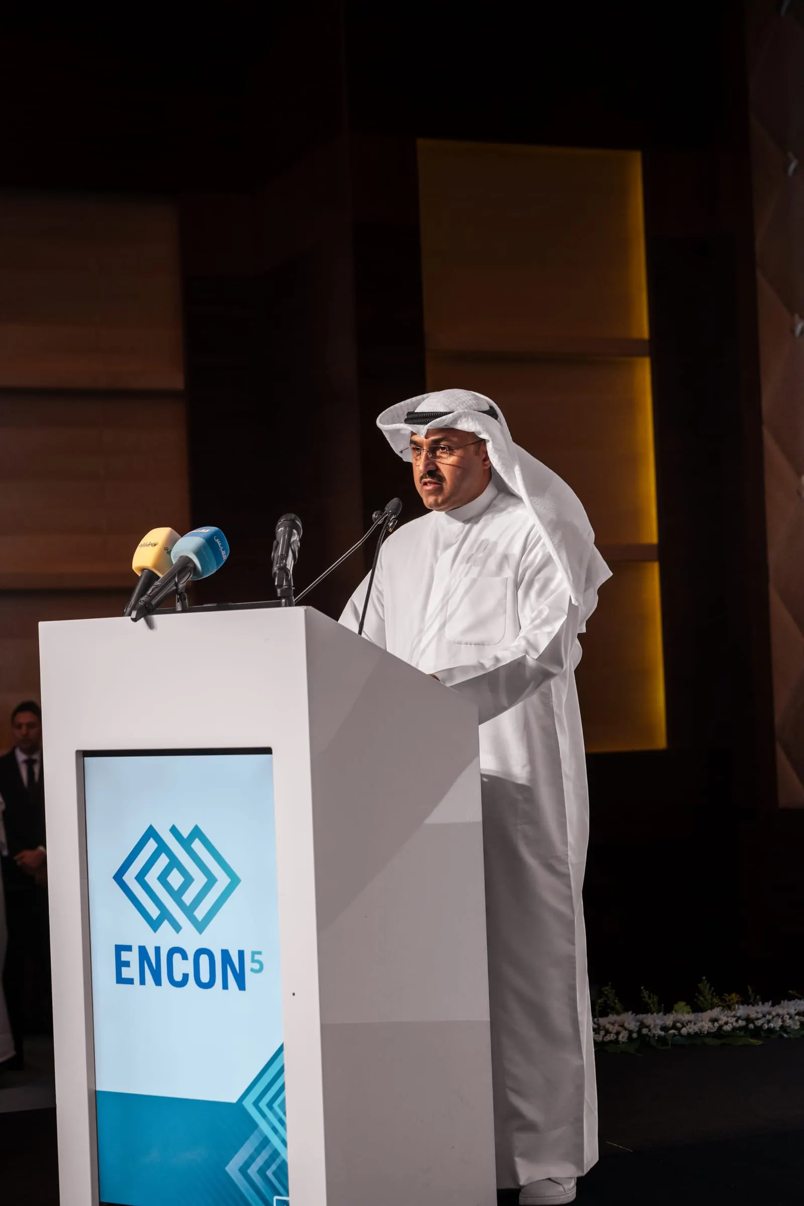 A speaker in Kuwaiti dress addresses the forum from an ENCON 5 podium with media microphones.