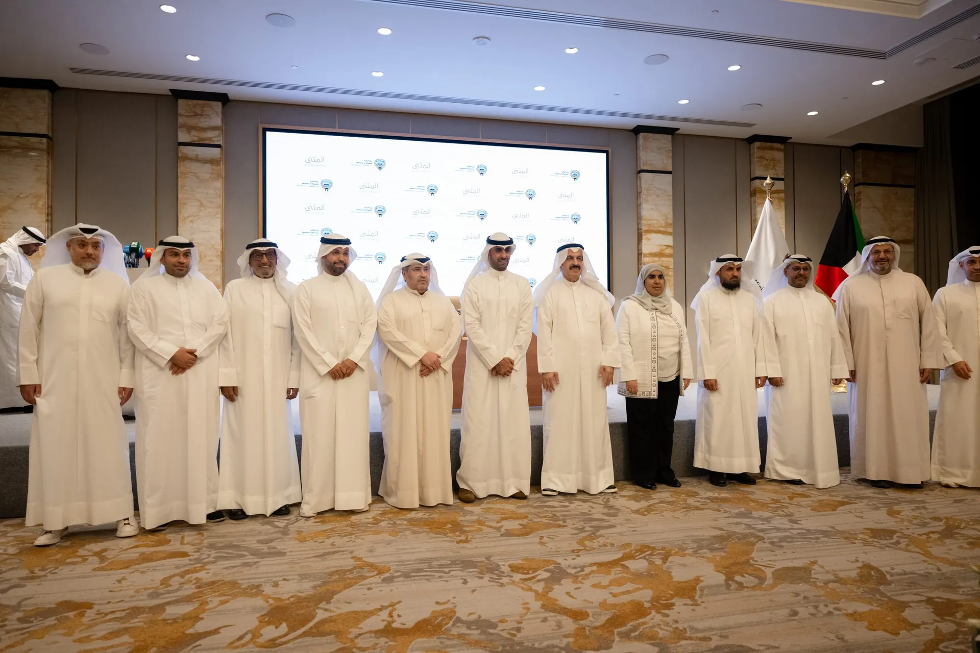 Twelve guests pose for a group photo in front of a screen repeating the Al Muthanna and Ministry of Finance logos, beside the Kuwaiti flag.