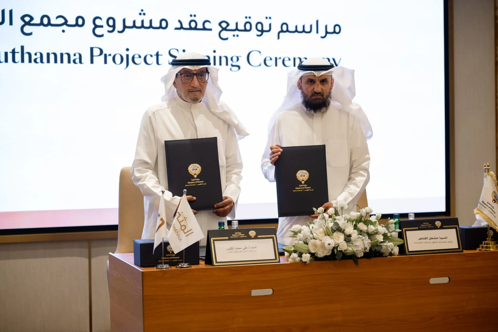Two signatories stand behind the signing table holding Ministry of Finance contract folders in front of an "Al Muthanna Project Signing Ceremony" screen.