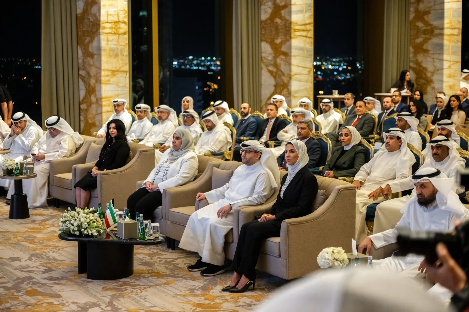 Guests in armchairs and rows of chairs watch the ceremony in a hall with marble columns and a night city view, with a camera operator in the foreground.