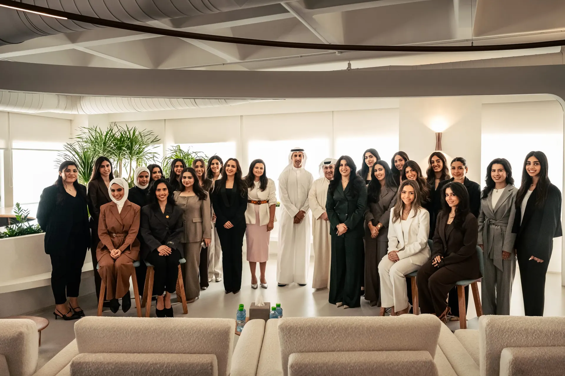 About thirty programme participants, mostly young women in business attire, pose with two men in Kuwaiti dress in a bright office lounge.