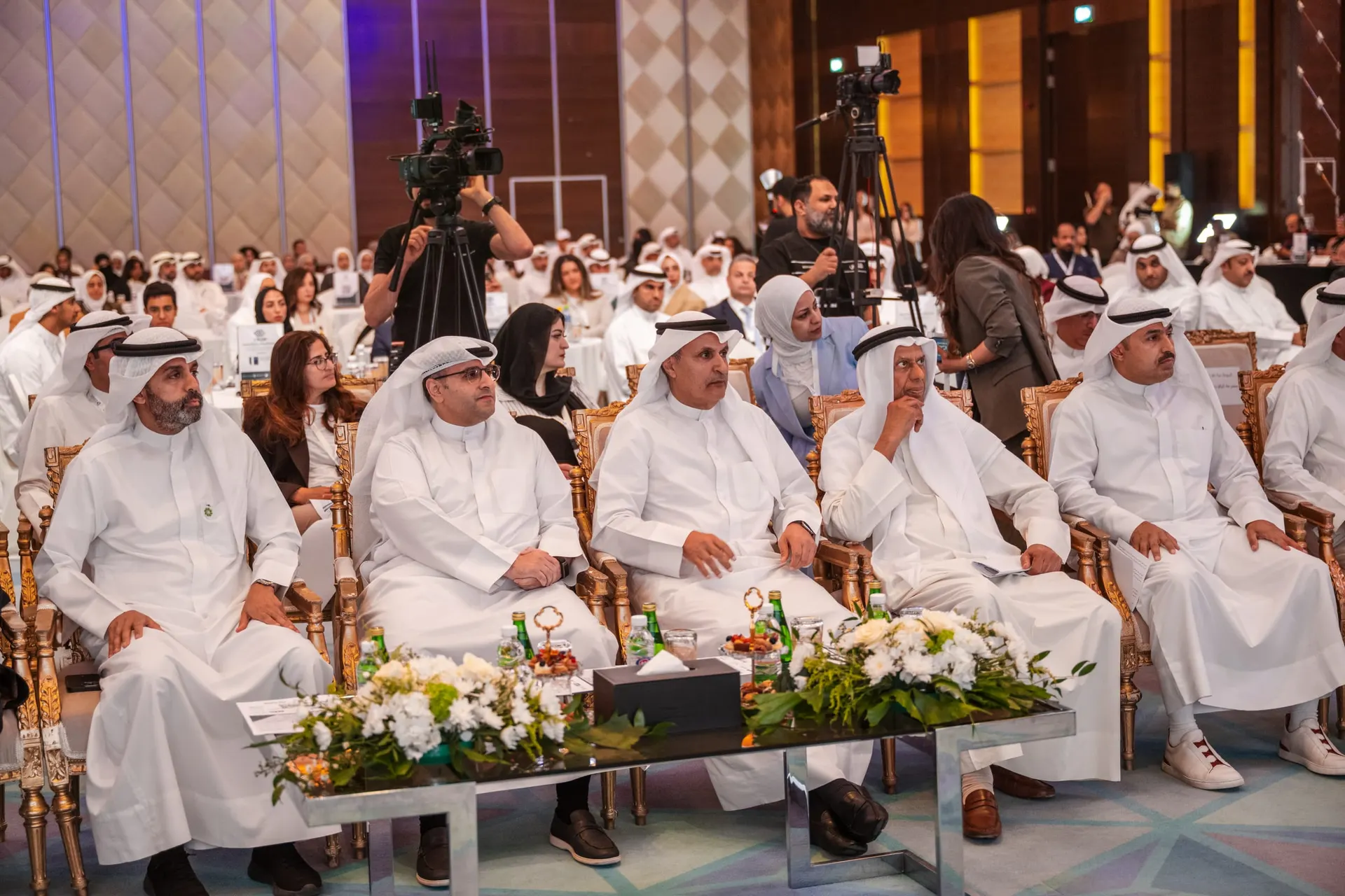 Senior guests sit in the front row of a large conference hall as camera operators film and a full audience fills the tables behind them.