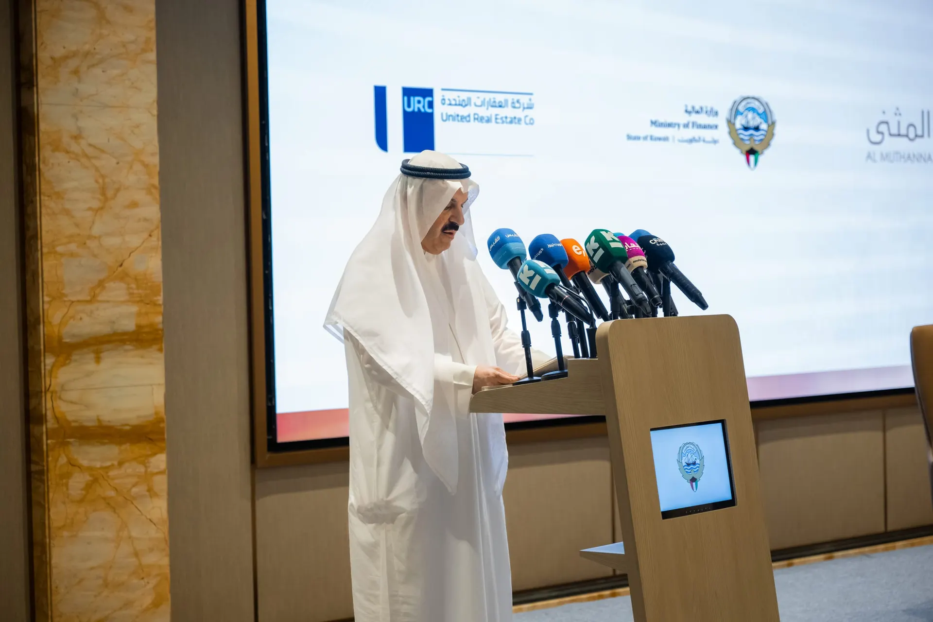 A speaker in Kuwaiti dress reads a statement at a podium lined with media microphones, in front of the URC, Ministry of Finance and Al Muthanna logos.