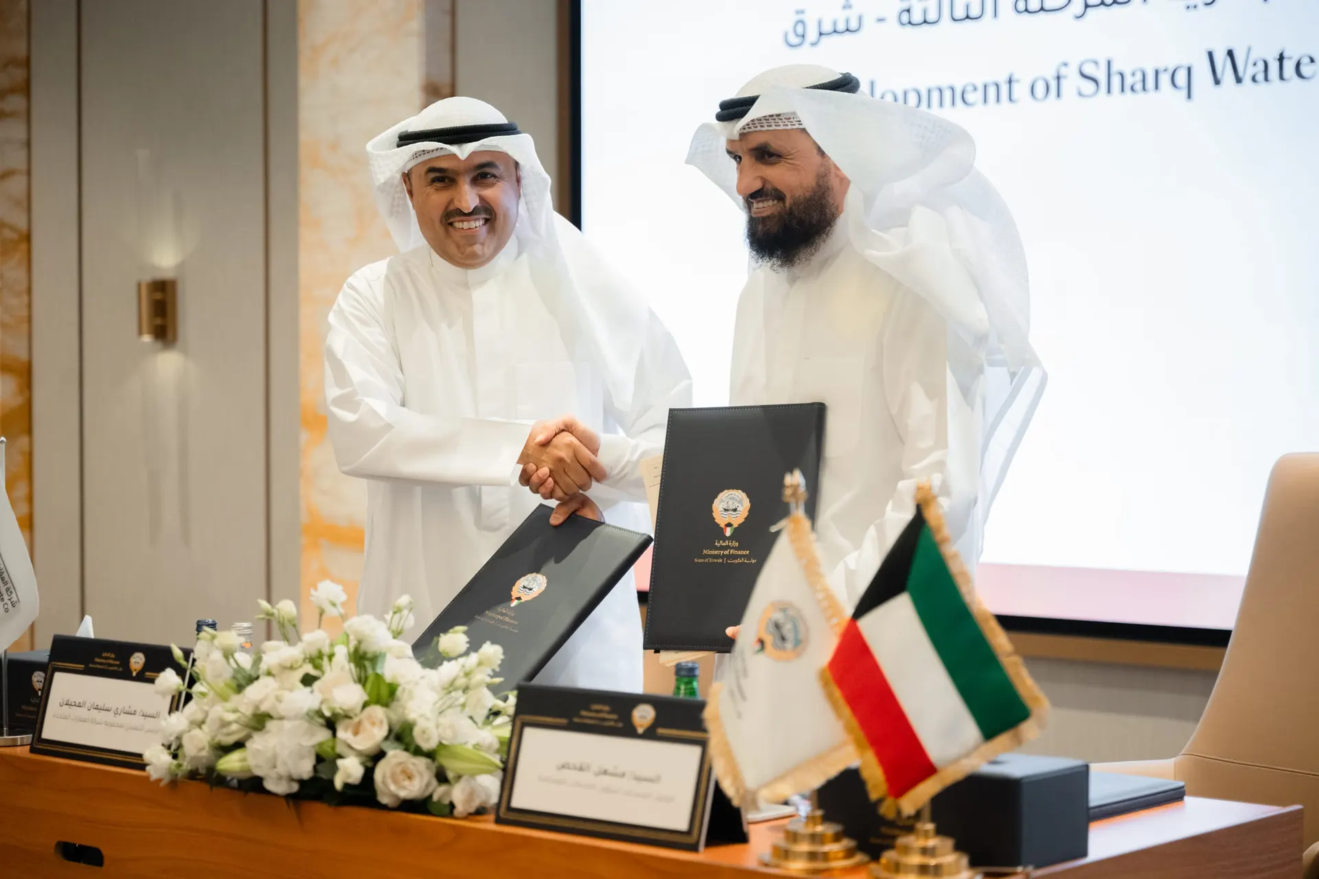 Two men in Kuwaiti dress shake hands and hold Ministry of Finance contract folders in front of a "Development of Sharq Waterfront" screen.