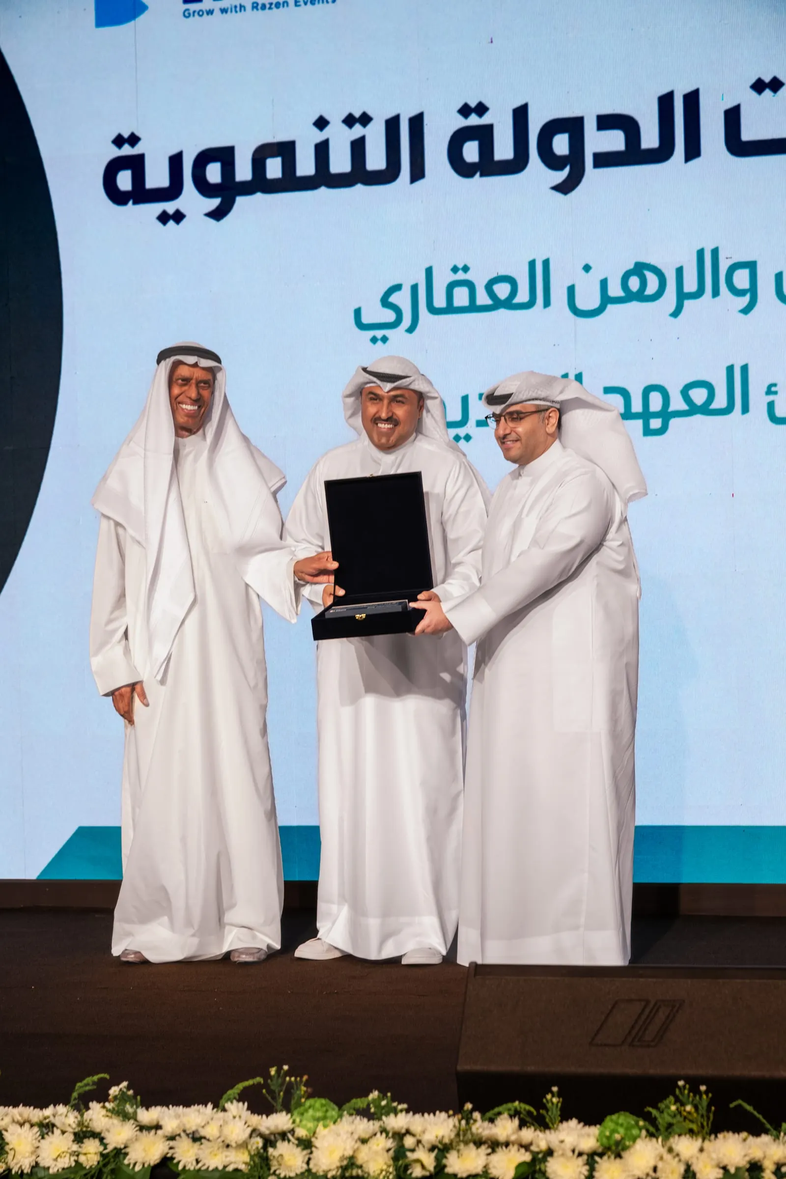 Three men in Kuwaiti dress present a commemorative plaque on stage in front of the forum screen.