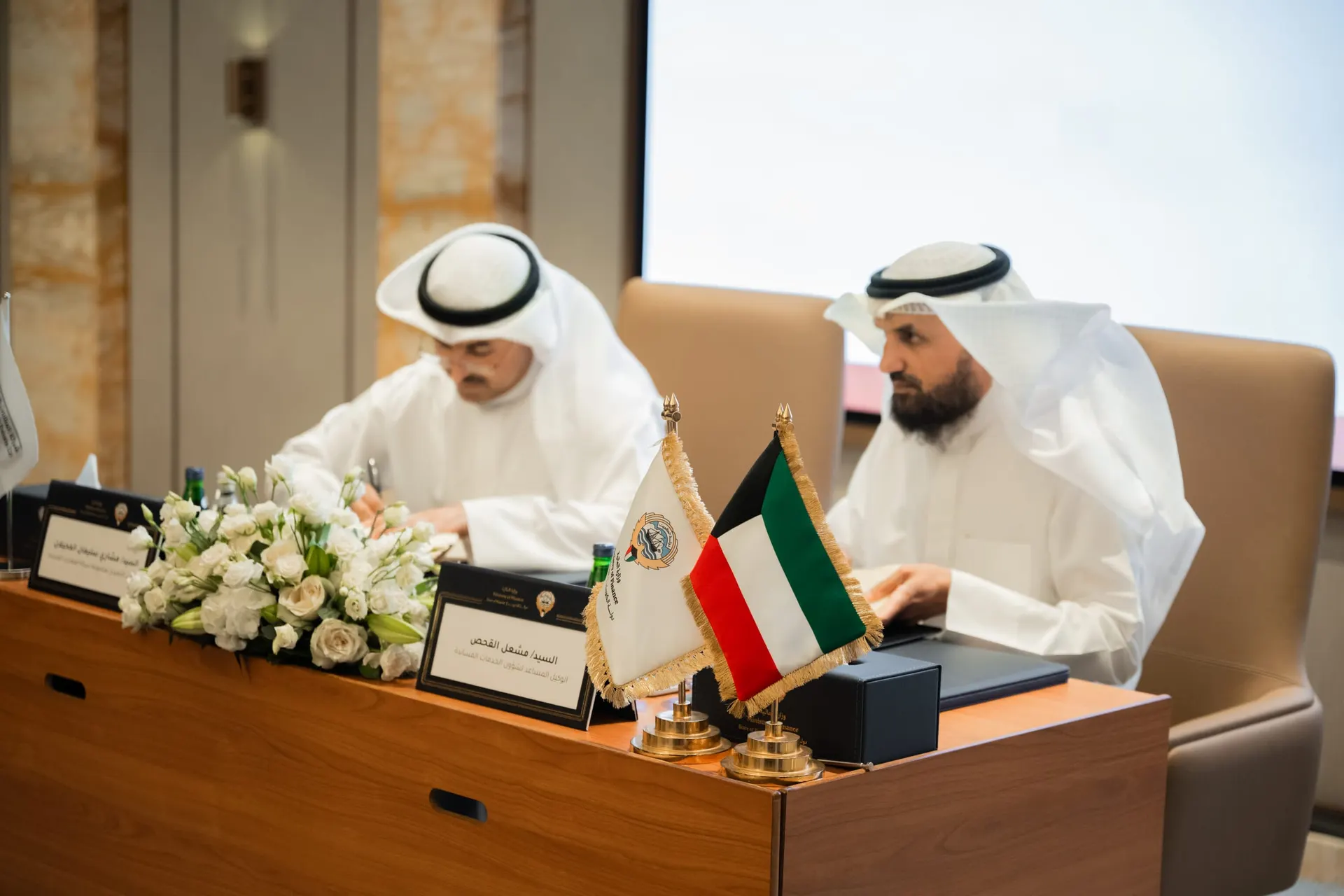 Two officials sign contracts at a table set with white flowers, name cards, the Kuwaiti flag and the Ministry of Finance flag.