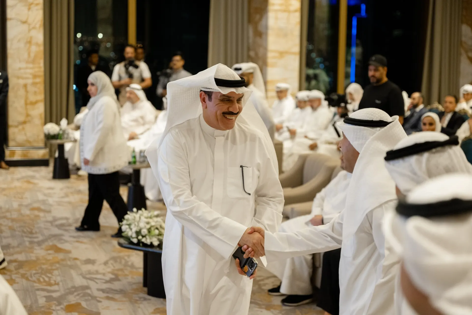 A smiling guest in Kuwaiti dress shakes hands with a seated guest as the audience and camera crews look on.